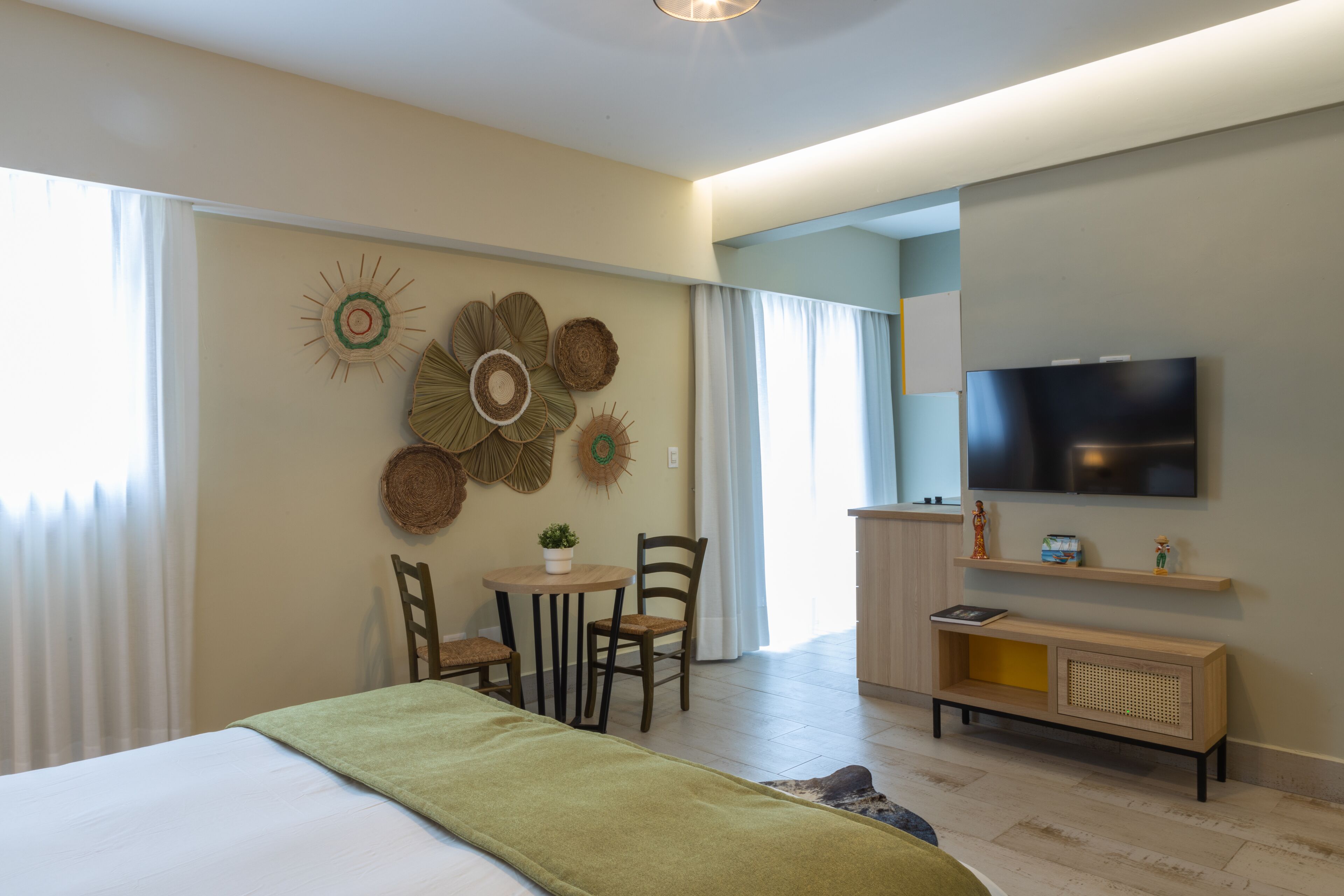 A modern hotel room with a bed, a dining area with chairs and a table, a television mounted on the wall, and decorative wall art.