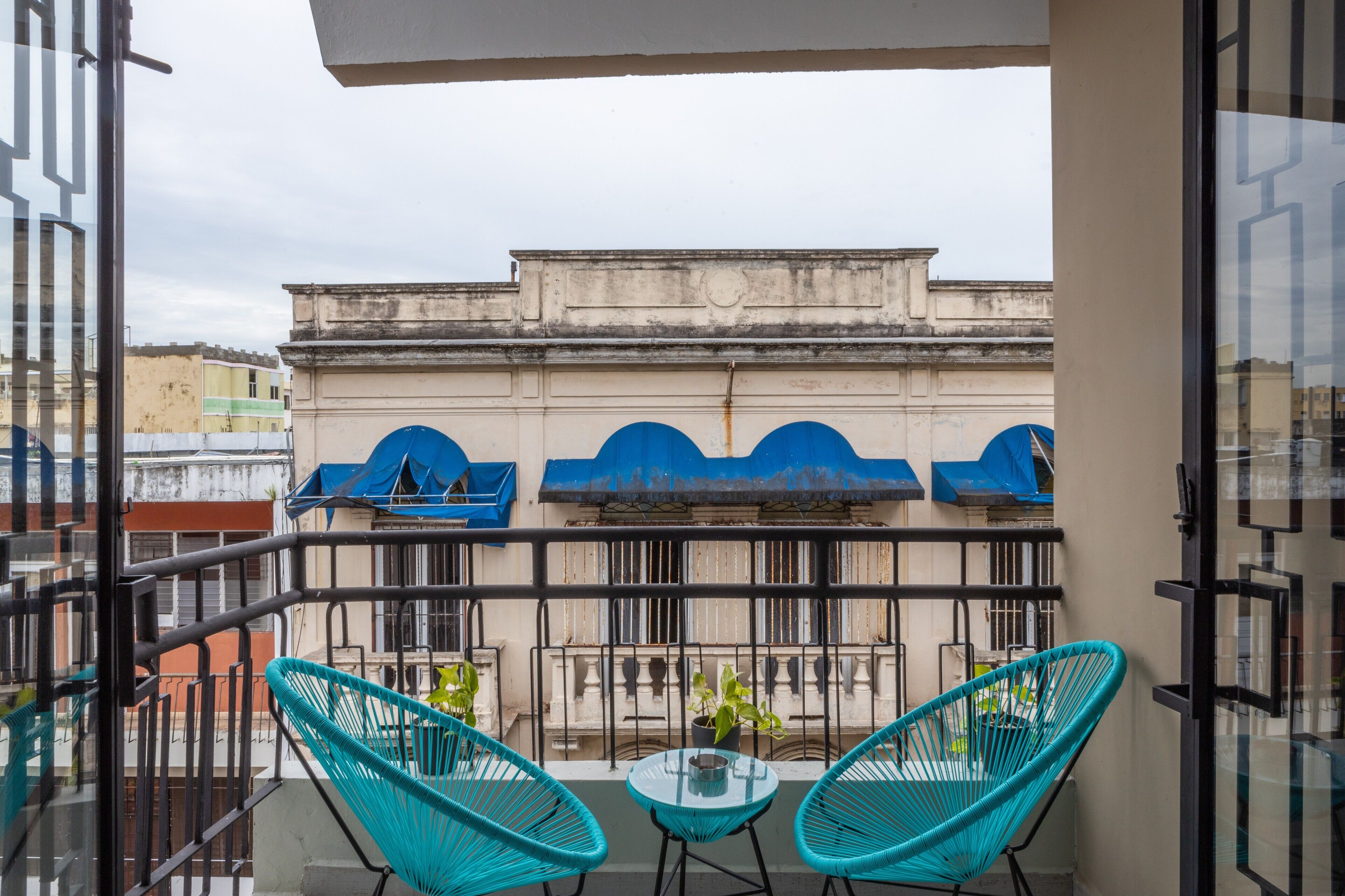 Ein Balkon mit zwei türkisfarbenen Stühlen und einem kleinen Tisch, mit Blick auf ein Gebäude mit blauen Markisen und einer beigen Fassade.
