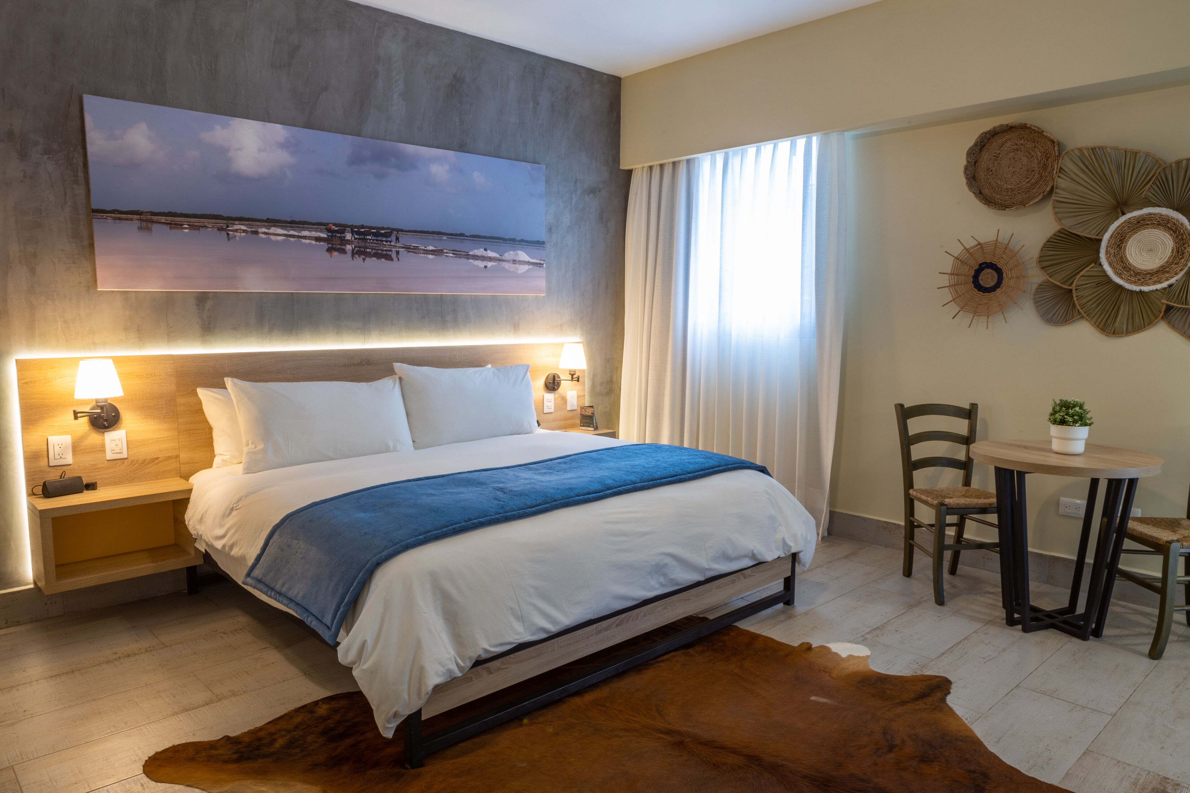 Een moderne hotelkamer met een groot bed, bedlampjes, uitzicht op zee, een klein tafeltje met stoelen, en een decoratief kunstwerk aan de muur.