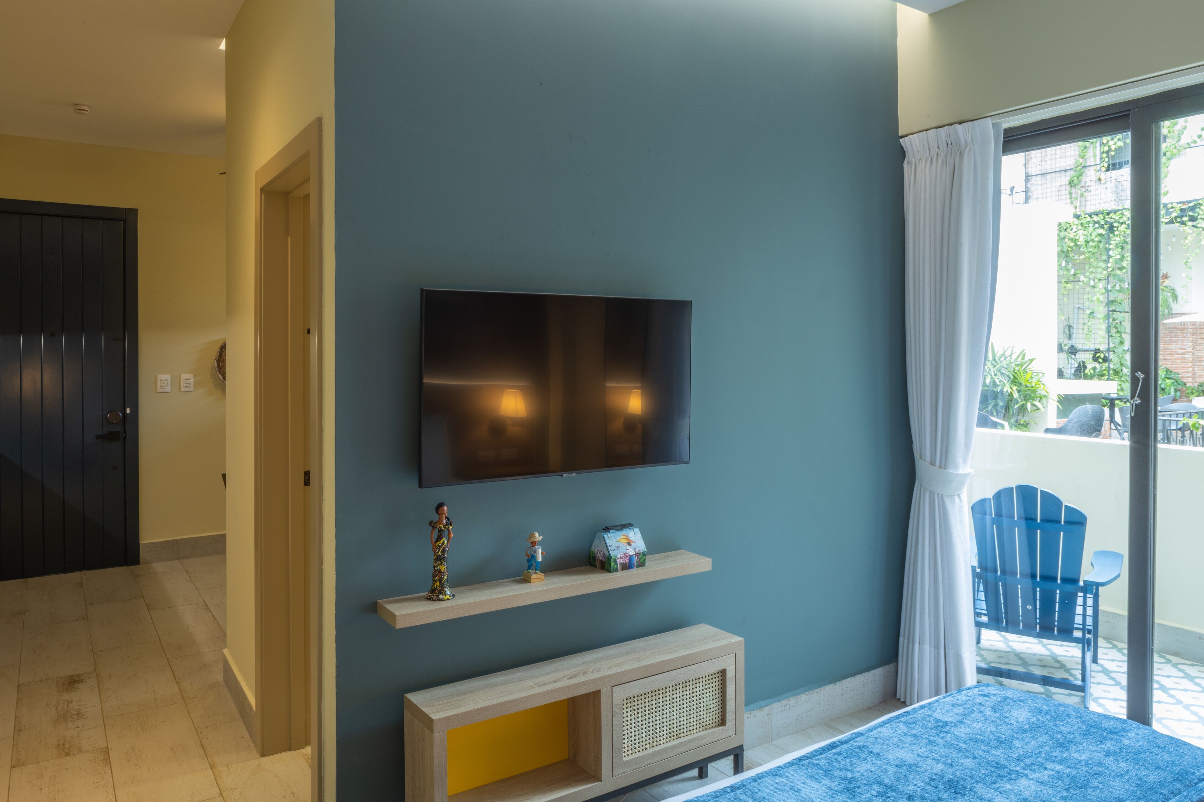 A room with a TV mounted on the wall, a small shelf with decorative items, a window with curtains, and a door leading to another room.
