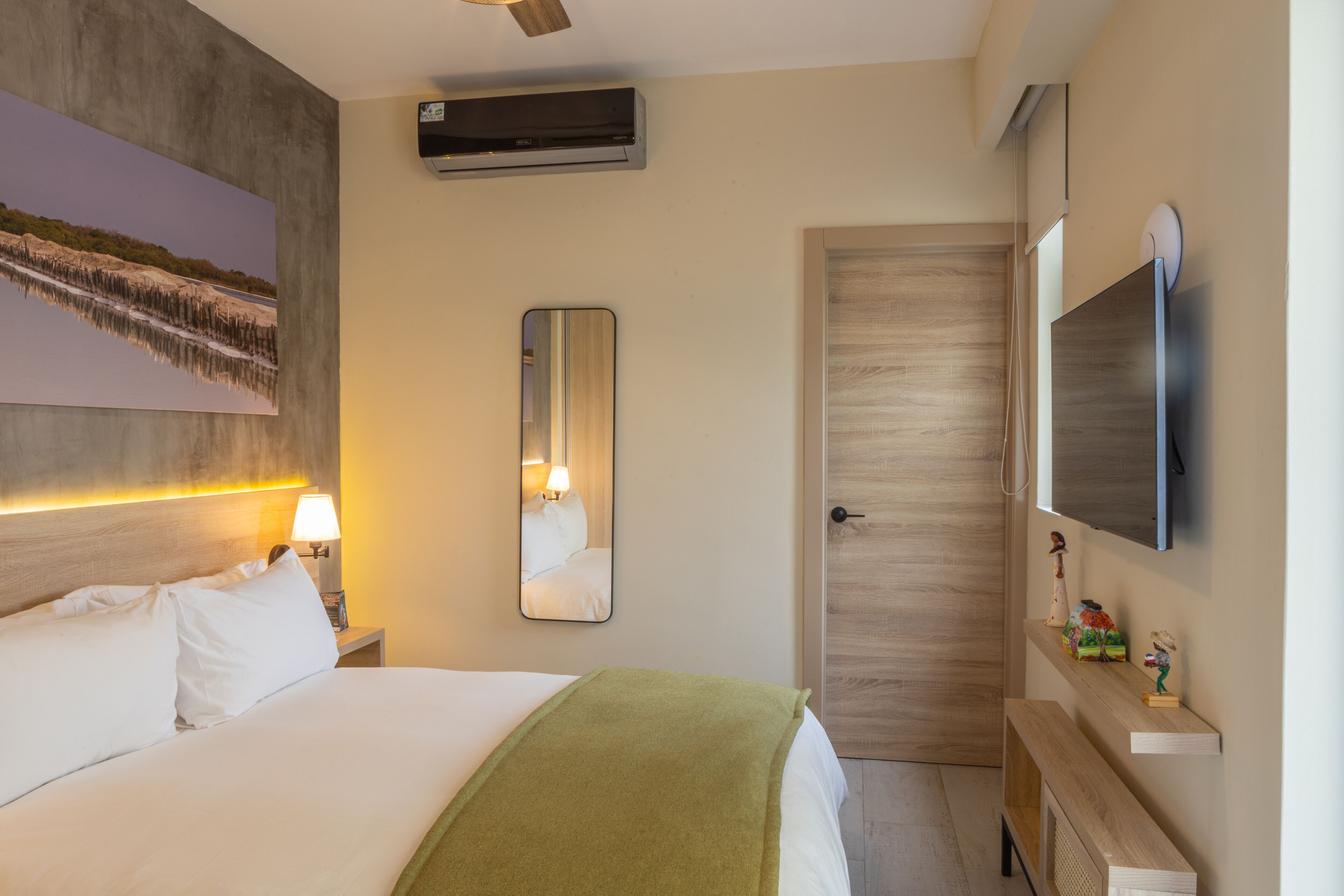 A hotel room with a bed, a bedside lamp, a mirror, a TV, and a wooden door.
