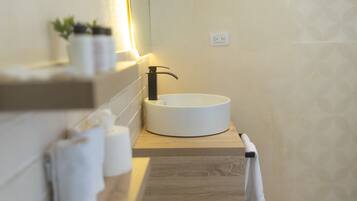 A bathroom with a round white sink, a black faucet, and a wooden countertop. There are two rolls of toilet paper and a potted plant on the shelf above the sink.