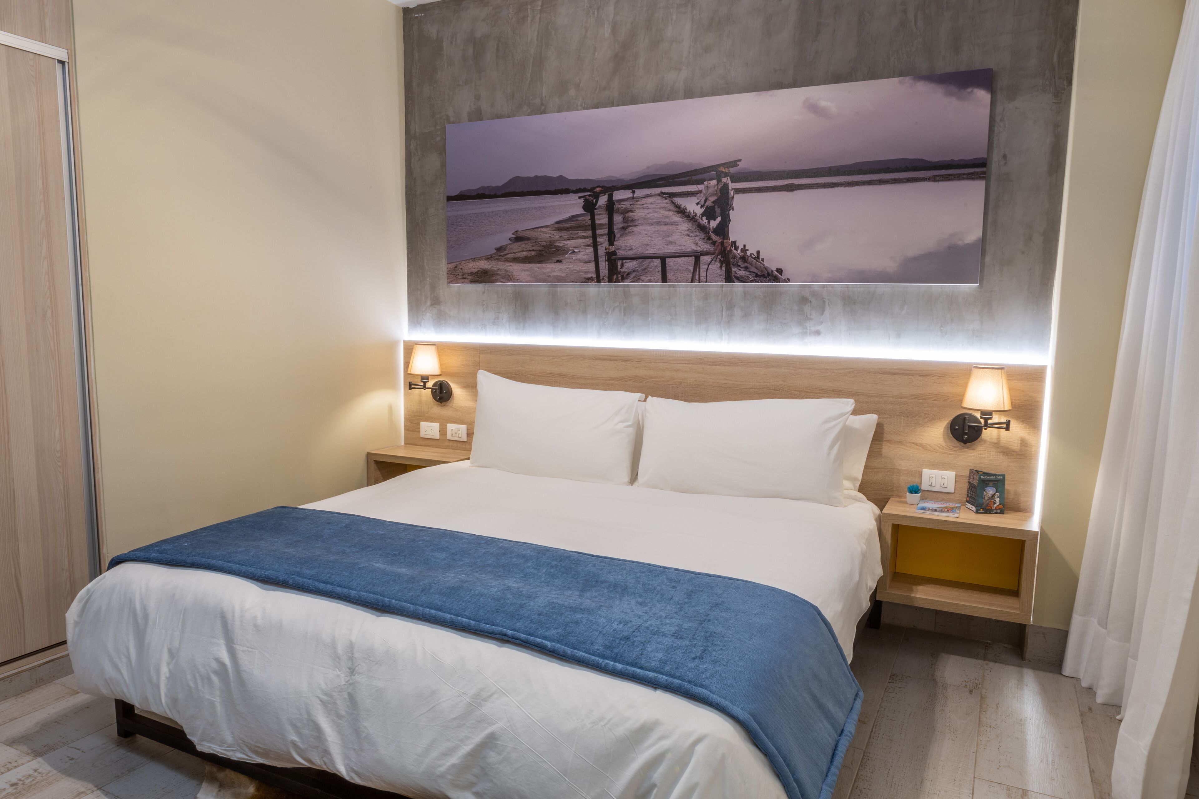 Ein Hotelzimmer mit Bett, Nachttisch, Wandlampe und einem Bild von einer Strandlandschaft.