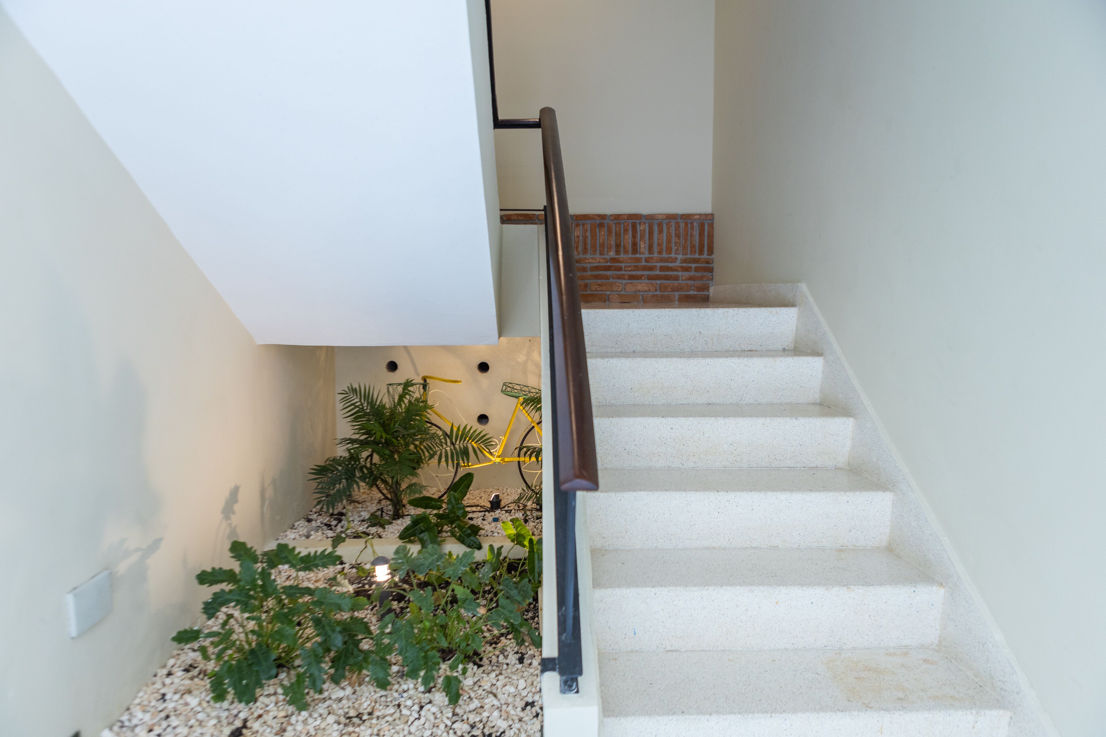 Una escalera con peldaños blancos y barandal de madera, que lleva a un rellano con pared blanca y zócalo de ladrillo.