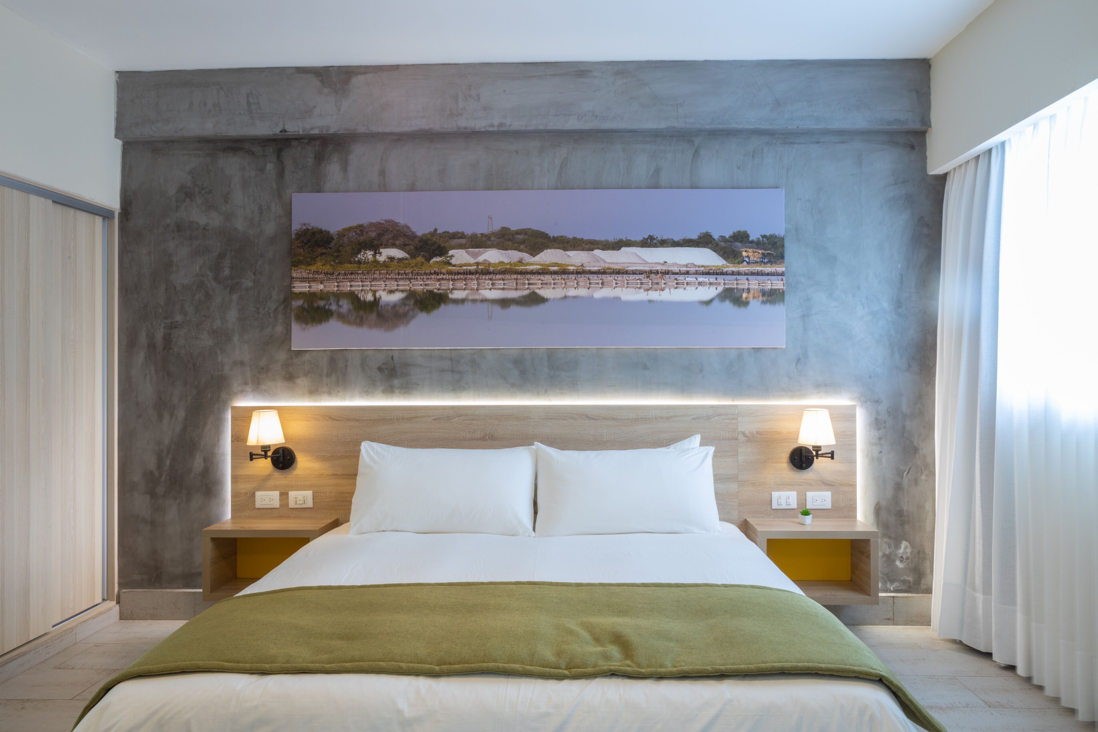 Habitación de hotel con una cama grande, dos mesitas de noche con lámparas, un cuadro sobre la cama y vistas a una masa de agua a través de la ventana.