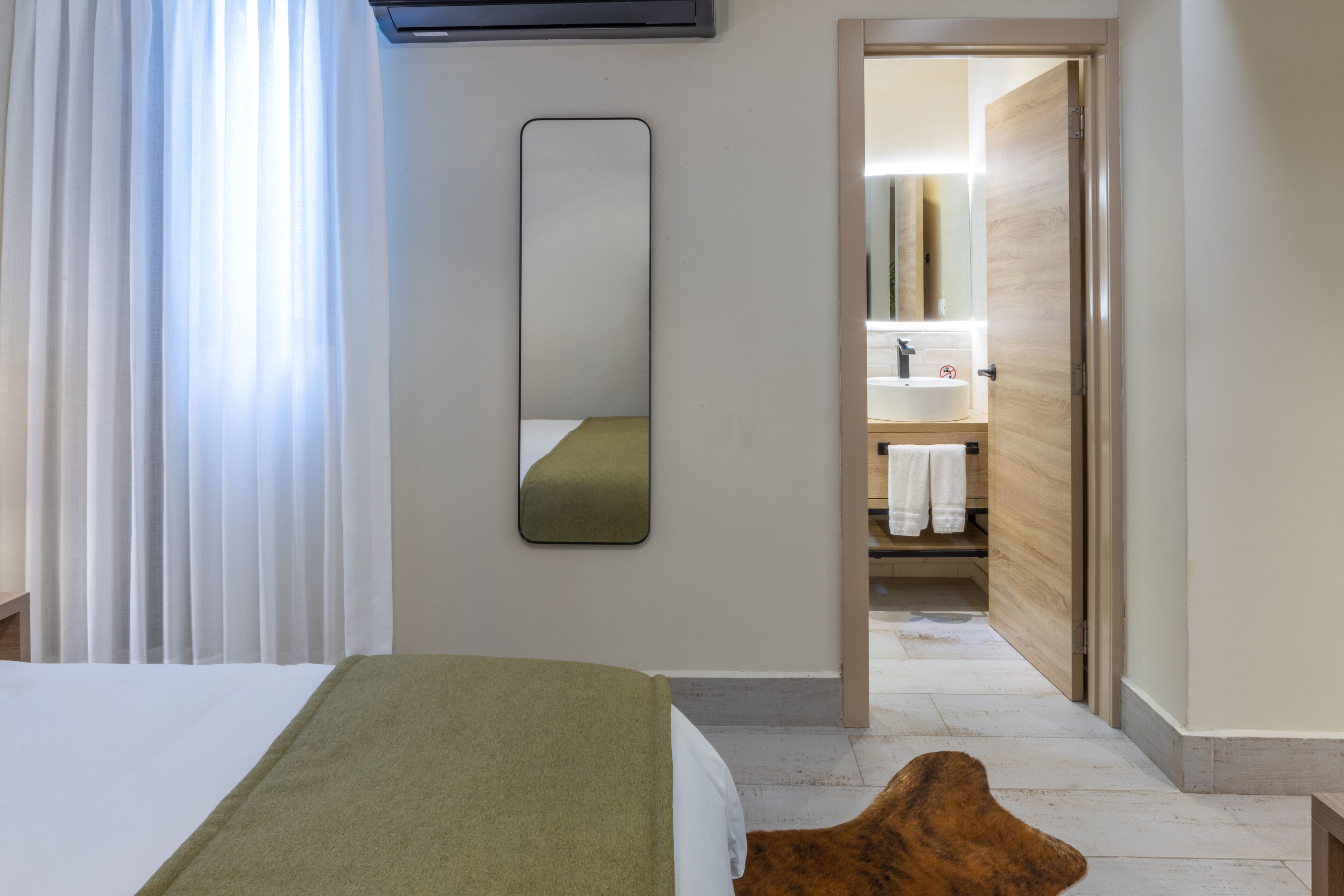 A hotel room with a bed, a mirror, a wooden door leading to a bathroom, and a white curtain.