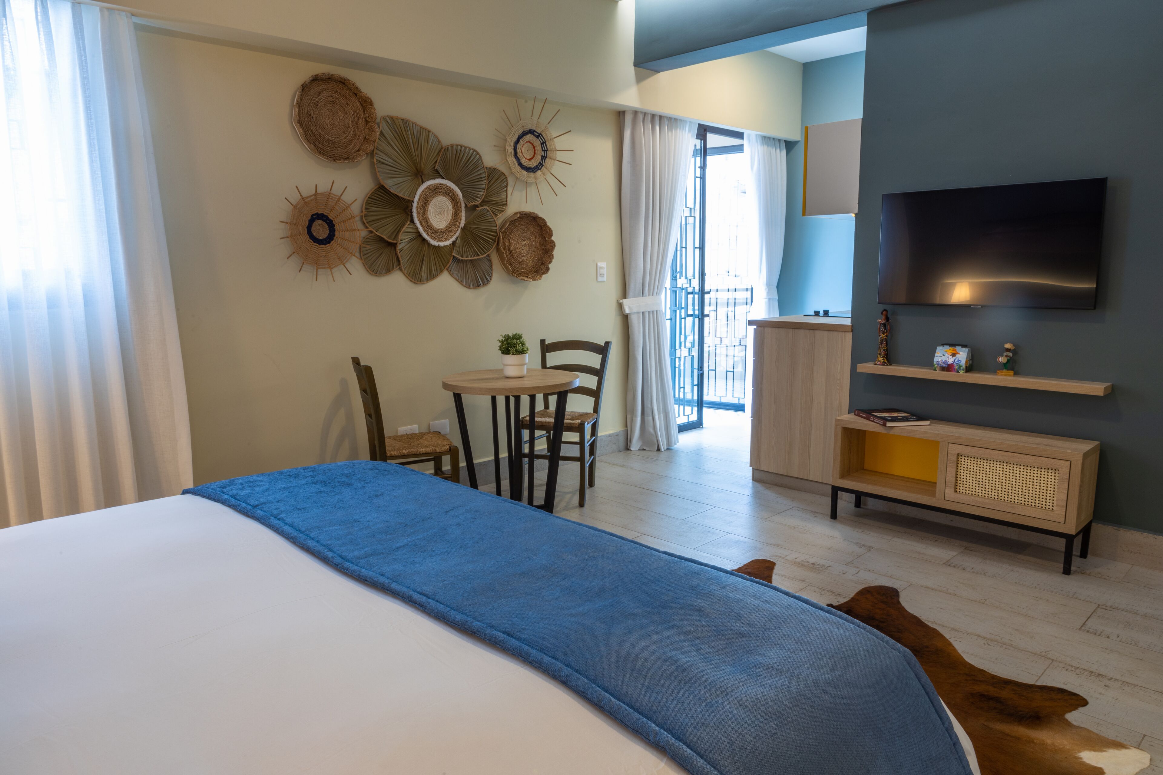 A hotel room with a bed, a desk, a chair, a TV, and a wall decorated with woven wall art.