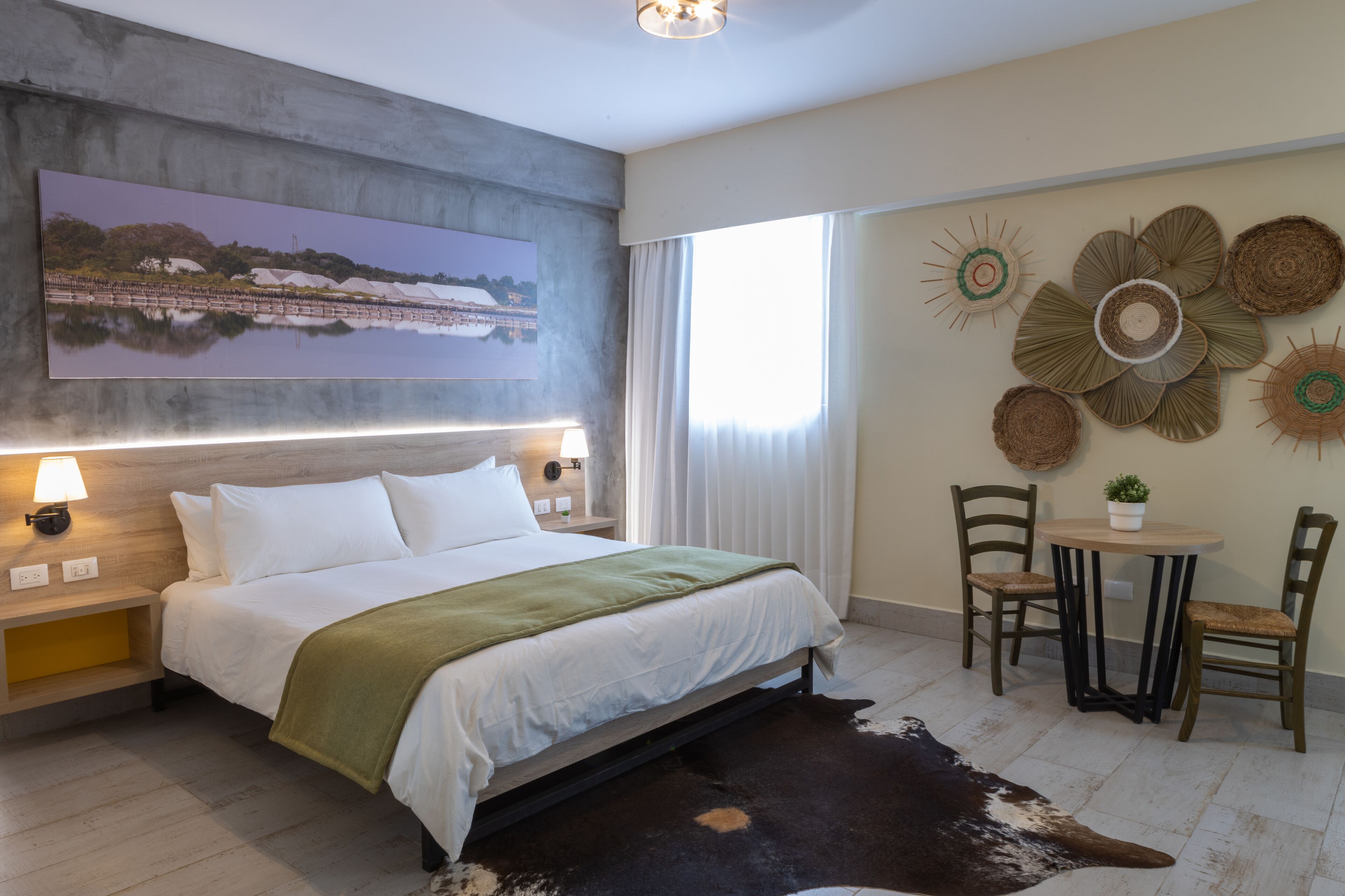 A modern hotel room with a large bed, a view of a lake, a dining area with a table and chairs, and decorative wall art.
