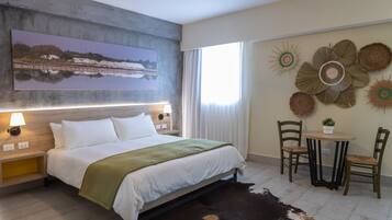 A modern hotel room with a large bed, a view of a lake, a dining area with a table and chairs, and decorative wall art.