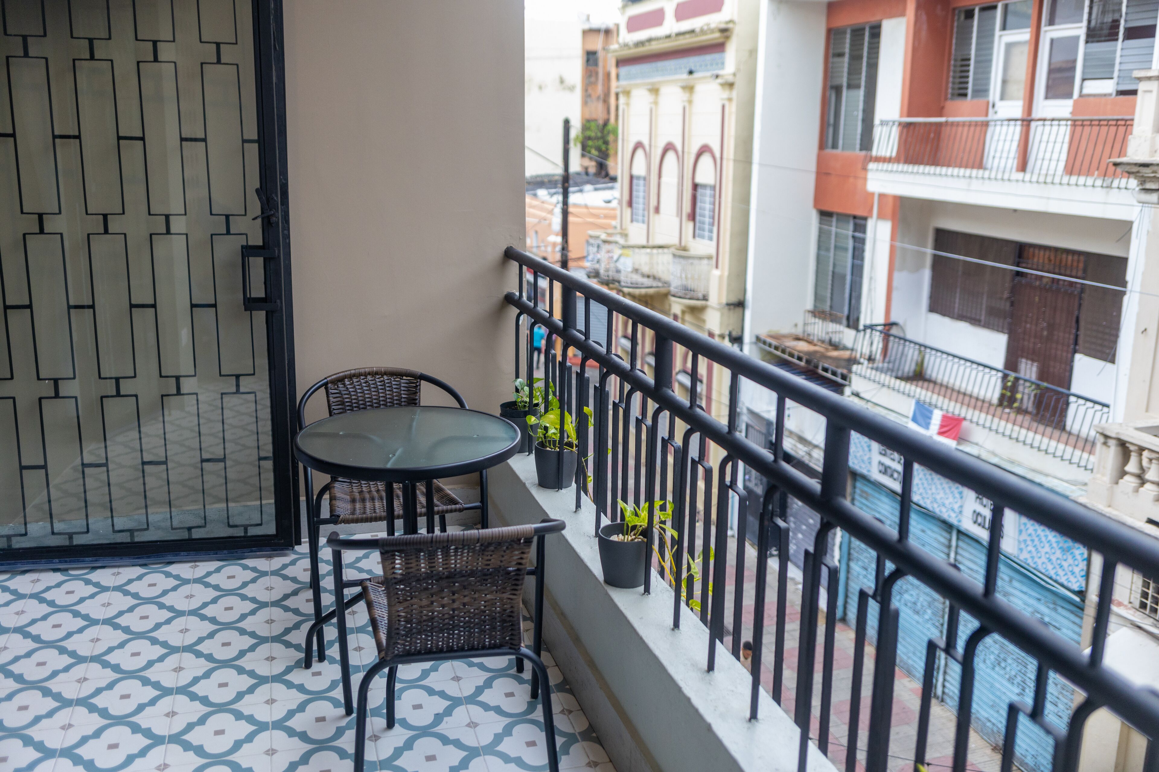 En balkon med et glasbord, fletstole, plantekrukker og udsigt til gaden nedenfor.