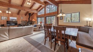Un vaste espace de vie doté d’un plafond en bois, de grandes fenêtres, d’une cheminée et d’une table à manger avec des chaises.
