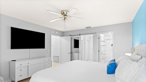 A bedroom with a large bed, a flat-screen TV mounted on the wall, a white dresser, and a ceiling fan.