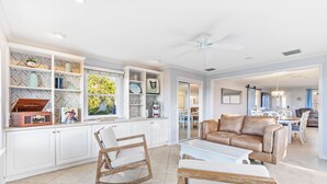 A spacious living room with a white sofa, a rocking chair, and a coffee table. There is a kitchen area with white cabinets and a herringbone-patterned backsplash. A dining area with a table and chairs is visible in the background.