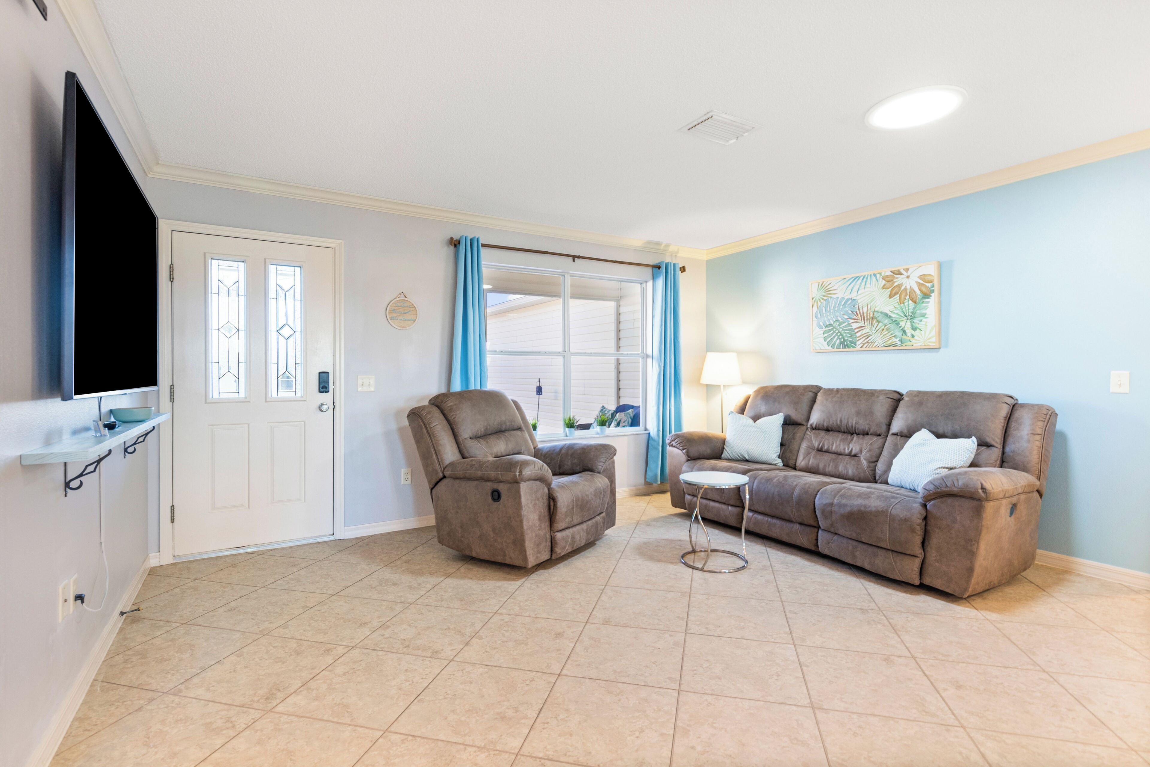 A living room with a beige tiled floor, a brown leather sofa, a recliner, a small round coffee table, a TV on a stand, a window with blue curtains, and a wall-mounted artwork.