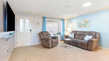 A living room with a beige tiled floor, a brown leather sofa, a recliner, a small round coffee table, a TV on a stand, a window with blue curtains, and a wall-mounted artwork.