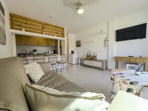 Uma sala de estar moderna com um sofá, um espaço de refeições com cadeiras, uma cozinha com armários de madeira, uma TV fixada na parede e um ventilador de teto.