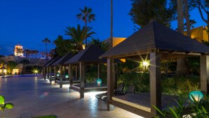 A series of covered outdoor seating areas with chairs and tables, surrounded by palm trees and a pool in the background. - Rosa Atlantic Apartment (Corralejo)