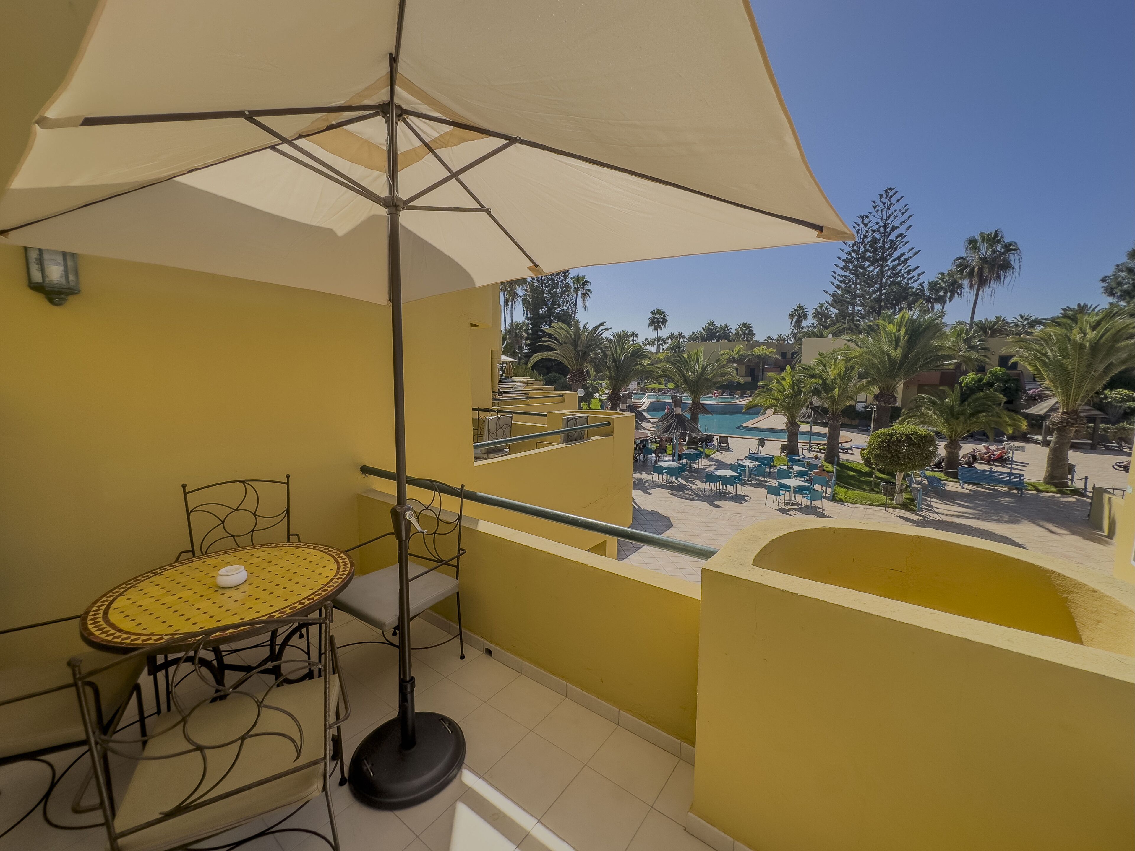Un balcon avec une table et une chaise, un grand parasol, et une vue sur un espace piscine agrémenté de palmiers et de chaises longues.