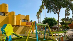 A playground with swings and slides, surrounded by palm trees and a building with yellow walls. - Rosa Atlantic Apartment (Corralejo)