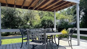 A covered outdoor deck with a wooden table and metal chairs, surrounded by a white fence and greenery.