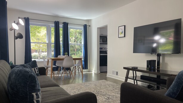 A modern living room with a sofa, a dining table with chairs, a TV, and a kitchen area visible through an open door.