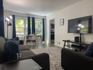 En moderne stue med sofa, spisebord med stole, fjernsyn og et køkken, der ses igennem en åben dør.