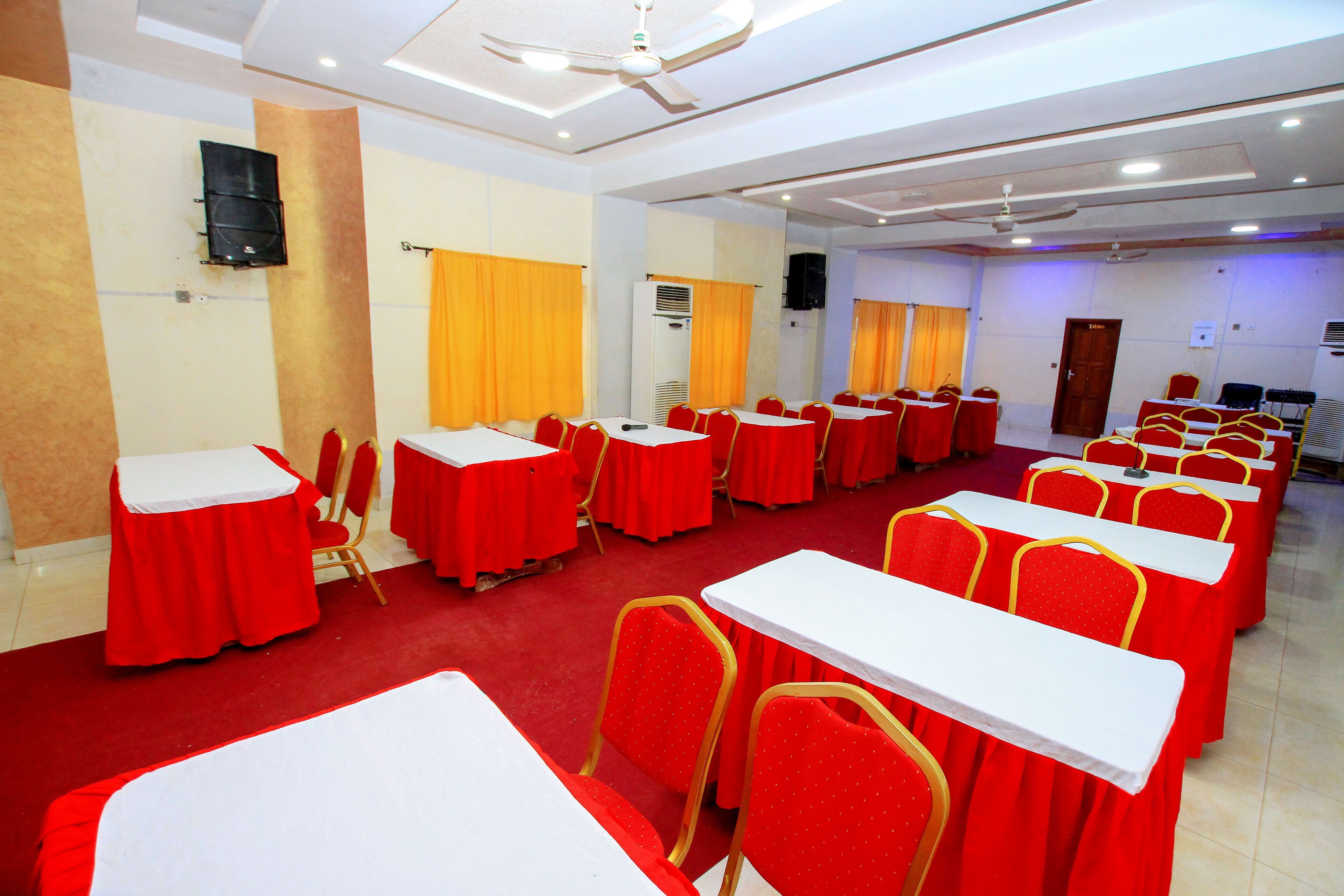 Un salón de conferencias con filas de mesas y sillas, ventiladores de techo y un equipo de aire acondicionado.