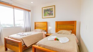 Een hotelkamer met twee houten bedden, een groot raam met gordijnen, een schilderij aan de muur en uitzicht op de stad.