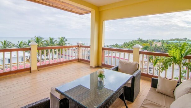 Een balkon met een glazen tafel en rieten meubilair, met uitzicht op een strand en palmbomen.