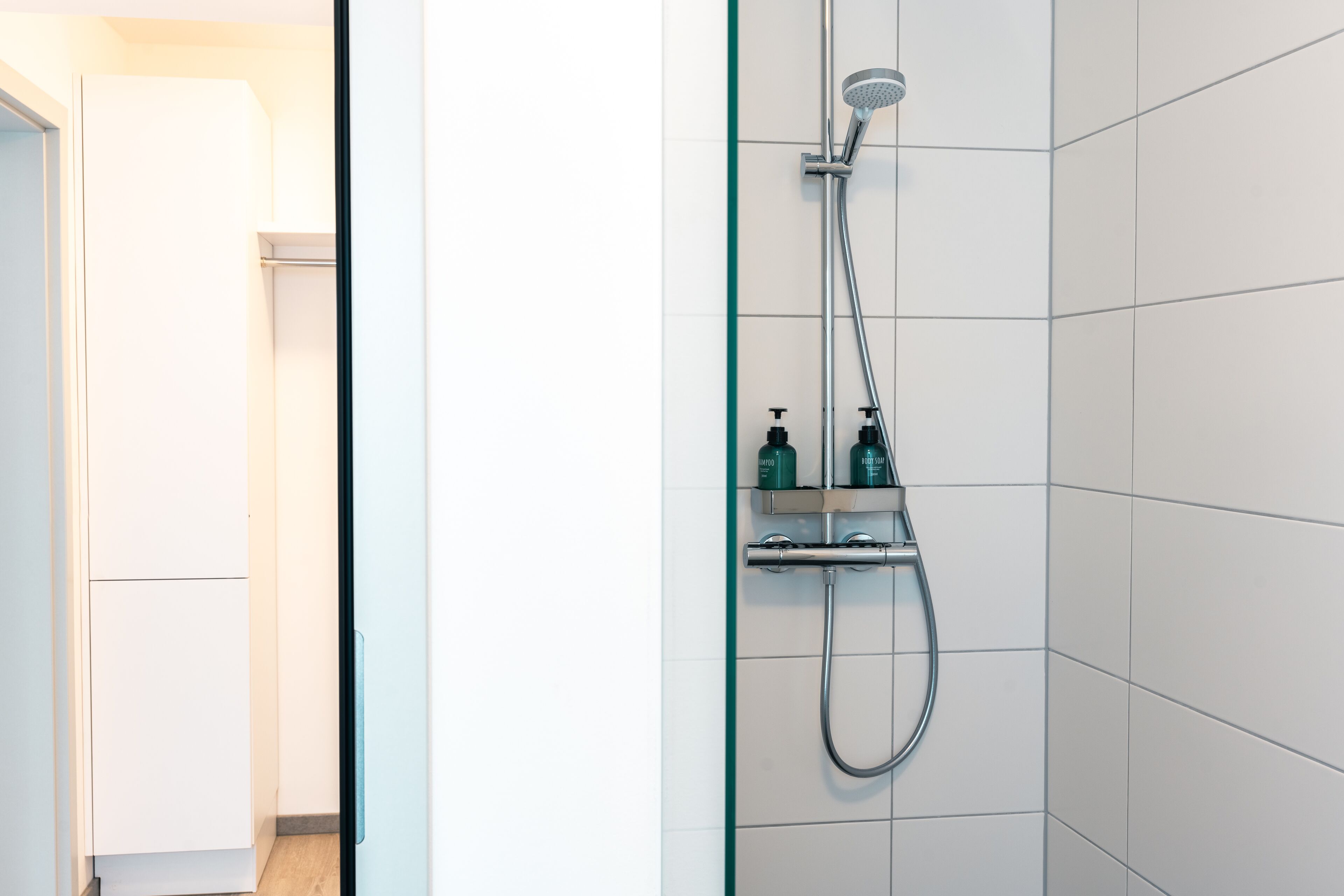 A modern bathroom with a glass shower enclosure, white tiles, and a shelf with toiletries.