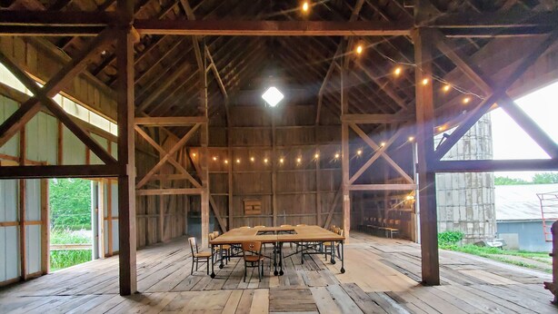 A rustic barn interior with a long wooden table and chairs, string lights, and a large window. - The Fox Den Comfy Lower Unit (Lake Elmo)