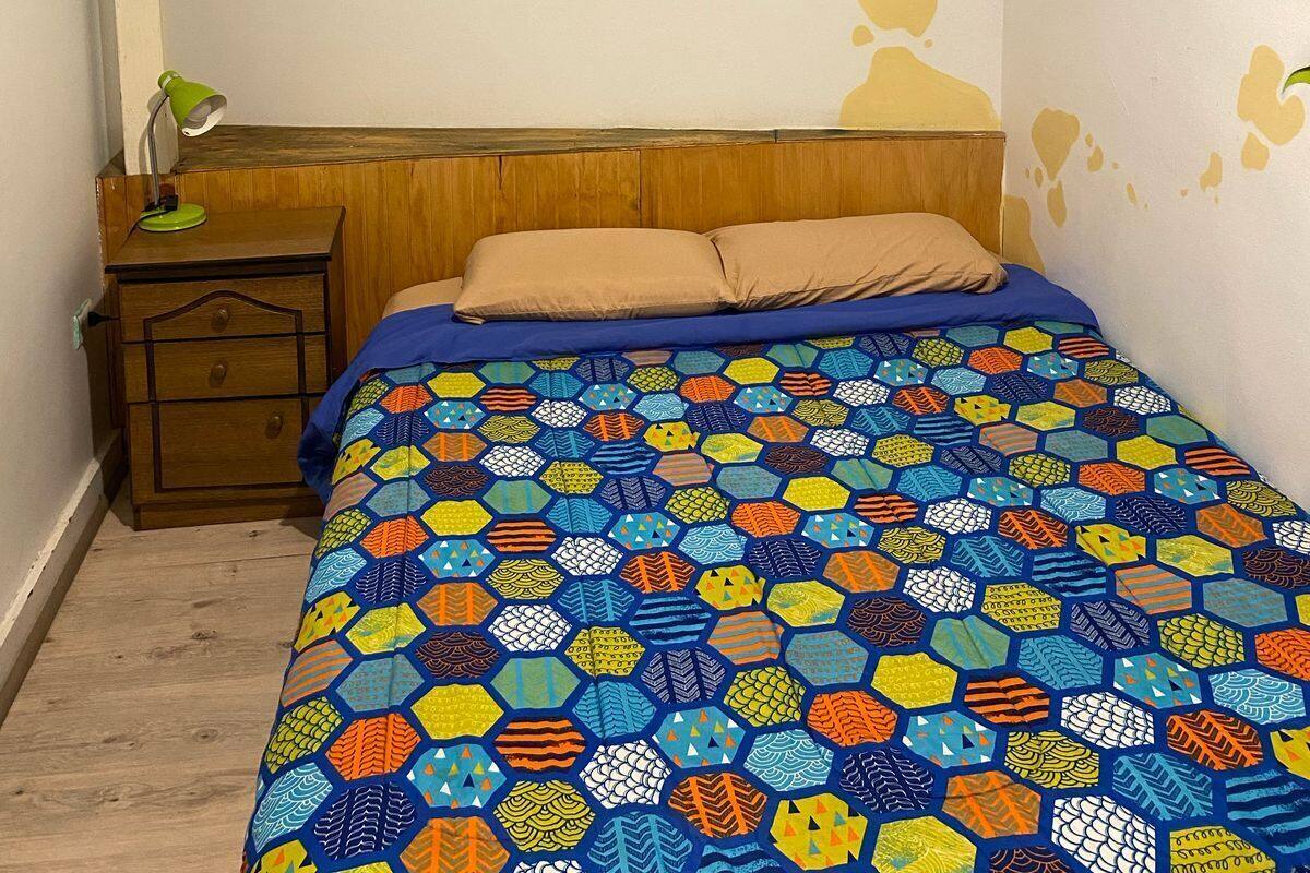 A bedroom with a large bed, a wooden headboard, a nightstand with a lamp, and a colorful hexagonal-patterned bedspread.
