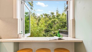 A kitchen counter with a window view of greenery, three wooden stools, and a magazine. - 2 bedroom tropical beach escape (Bondi Beach)