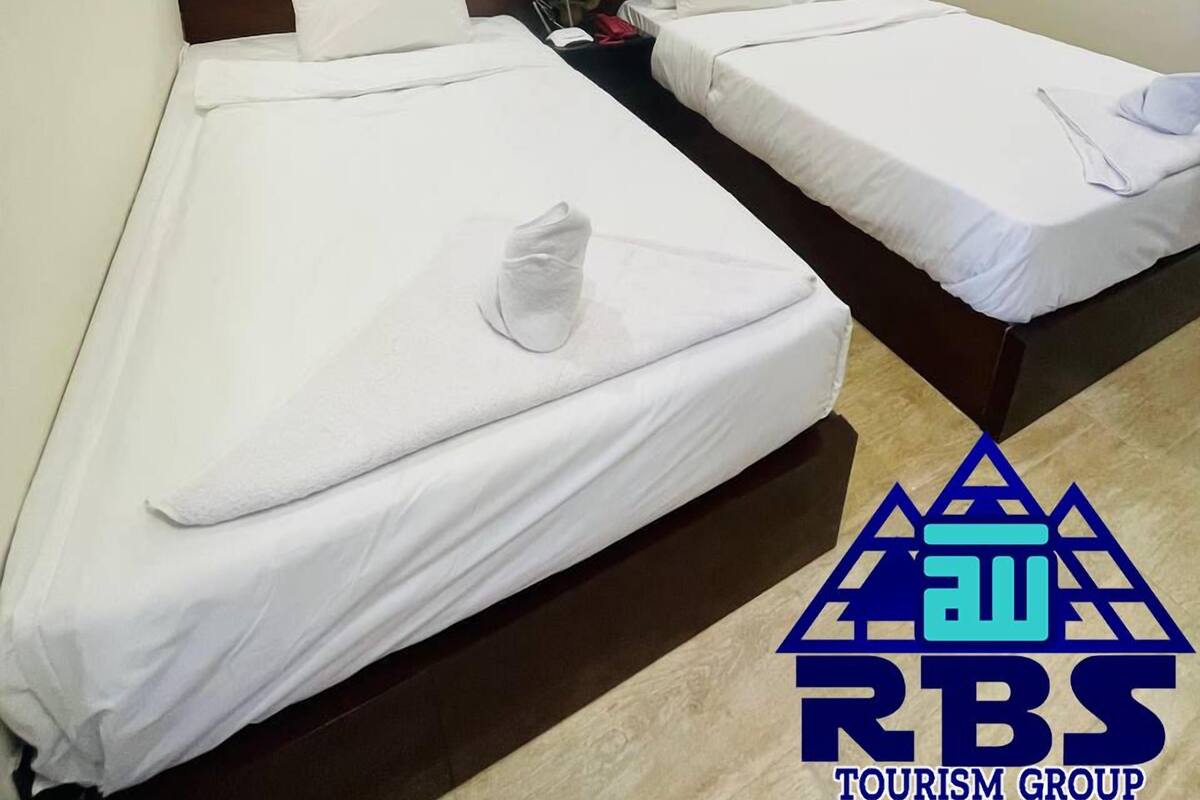 A hotel room with two single beds, white bedding, a nightstand with a vase of flowers, and a logo for RBS Tourism Group on the wall.