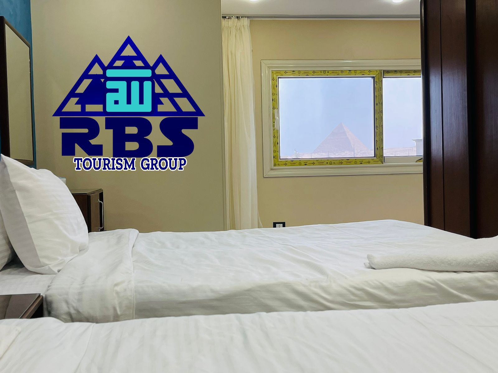 A hotel room with a bed, a window with a view of a pyramid, and a logo for RBS Tourism Group on the wall.