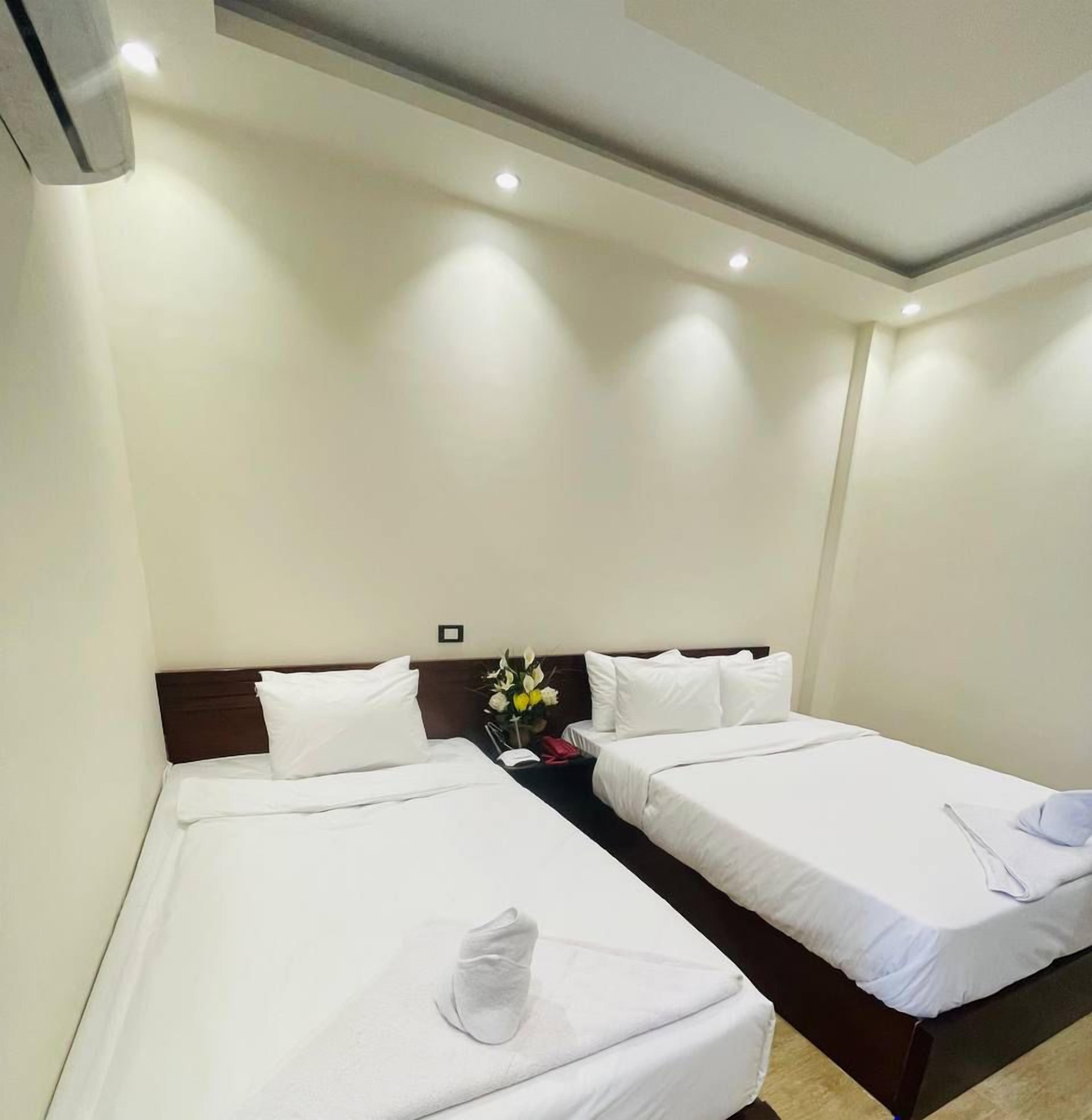 A hotel room with two single beds, white bedding, a nightstand with a vase of flowers, and a logo for RBS Tourism Group on the wall.