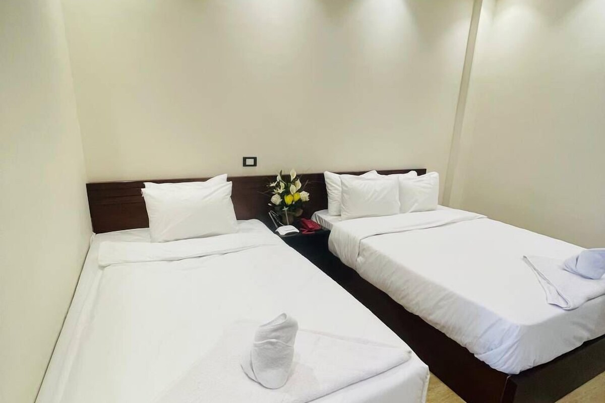 A hotel room with two single beds, white bedding, a nightstand with a vase of flowers, and a logo for RBS Tourism Group on the wall.