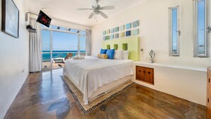 A bedroom with a large bed, a ceiling fan, a TV mounted on the wall, and a view of the ocean through a sliding door. - 4 BR Oceanfront Villa with Pool (Isla Mujeres)
