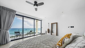 Une chambre dotée d’un grand lit, d’un ventilateur de plafond et offrant une vue sur l’océan grâce à de grandes fenêtres du sol au plafond.