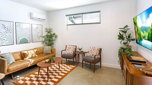 Una sala de estar moderna con un sofá, dos sillones, una mesa de centro y un televisor. Hay unidades de aire acondicionado instaladas en la pared y una ventana con persianas.