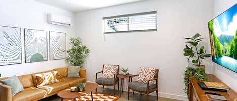 Una sala de estar moderna con un sofá, dos sillones, una mesa de centro y un televisor. Hay unidades de aire acondicionado instaladas en la pared y una ventana con persianas.