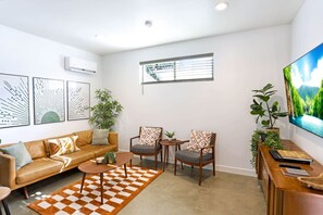 Un salon moderne comprenant un canapé, deux fauteuils, une table basse et un téléviseur. On y trouve également des climatiseurs fixés au mur et une fenêtre équipée de stores.