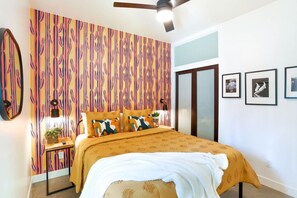 Une chambre à coucher avec un papier peint à motifs, un lit recouvert d’une housse de couette jaune, un ventilateur de plafond et des tableaux encadrés accrochés au mur.
