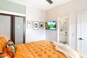 Un dormitorio con cama, televisor, puerta de baño y cuadros en la pared.