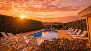 A swimming pool with lounge chairs, surrounded by hedges, during sunset.
