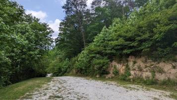 Um caminho de cascalho serpenteia por uma área arborizada, com vegetação densa e um declive rochoso.