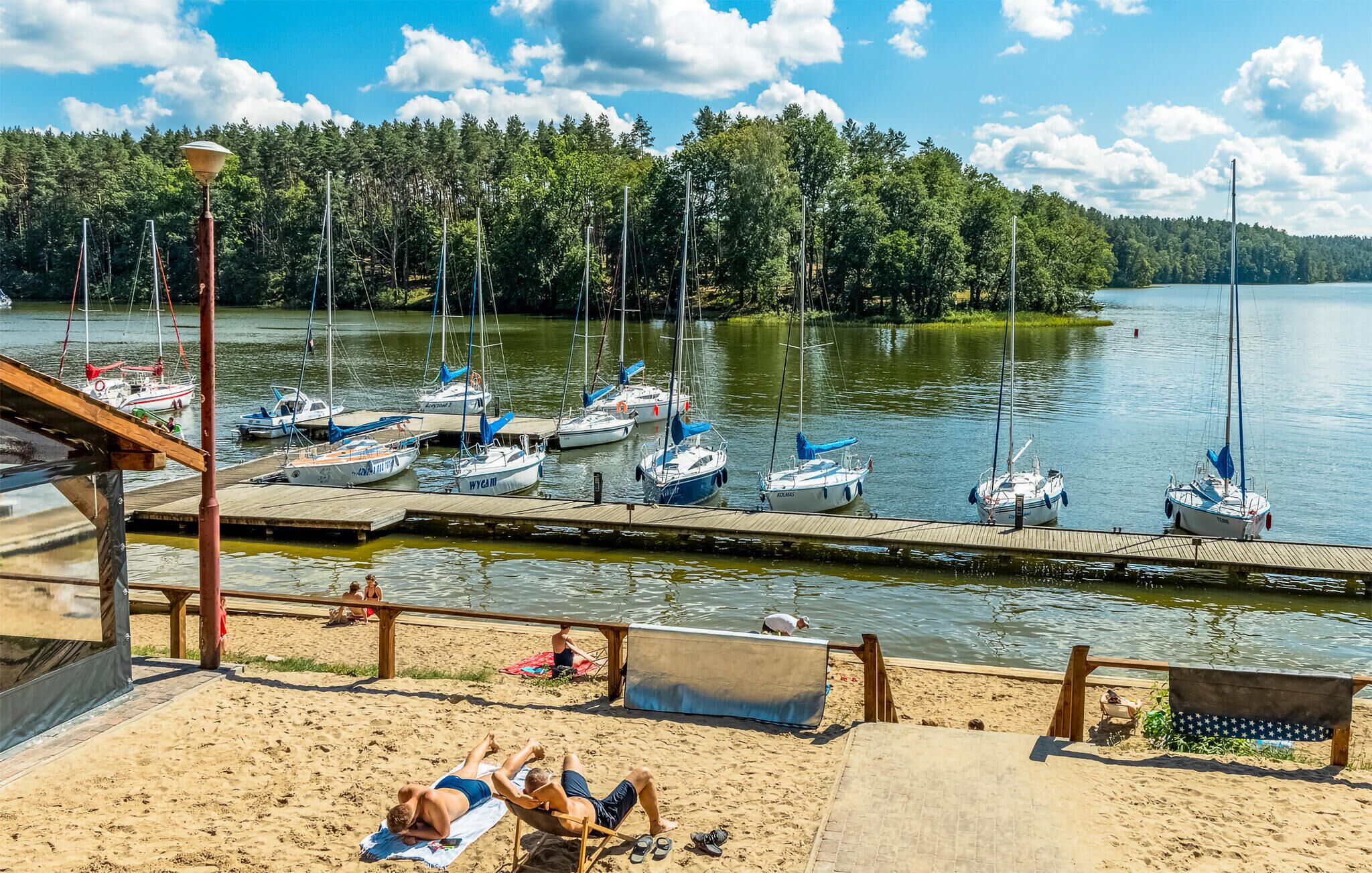 A lakeside dock with several boats, a sandy beach area with people sunbathing, and a forested shoreline in the background.