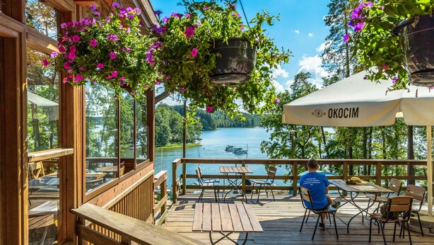 Eine Holzterrasse mit Tischen und Stühlen, die auf einen See blickt, mit hängenden Blumenkörben und einem überdachten Bereich mit einem Sonnenschirm, auf dem 'OKOCIM' steht.