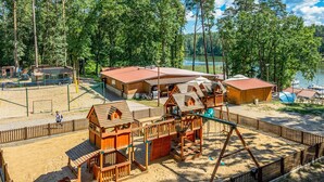 Ein Spielplatz mit Holzspielgeräten, einem Sandbereich und einem Gebäude mit rotem Dach.