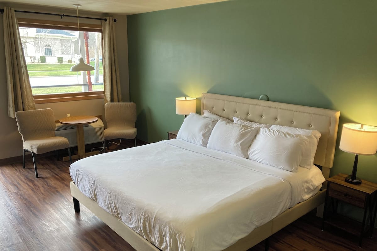 Ein Hotelzimmer mit einem großen Bett, zwei Nachttischen mit Lampen, einem Sessel und Blick auf ein Gebäude durch ein Fenster.